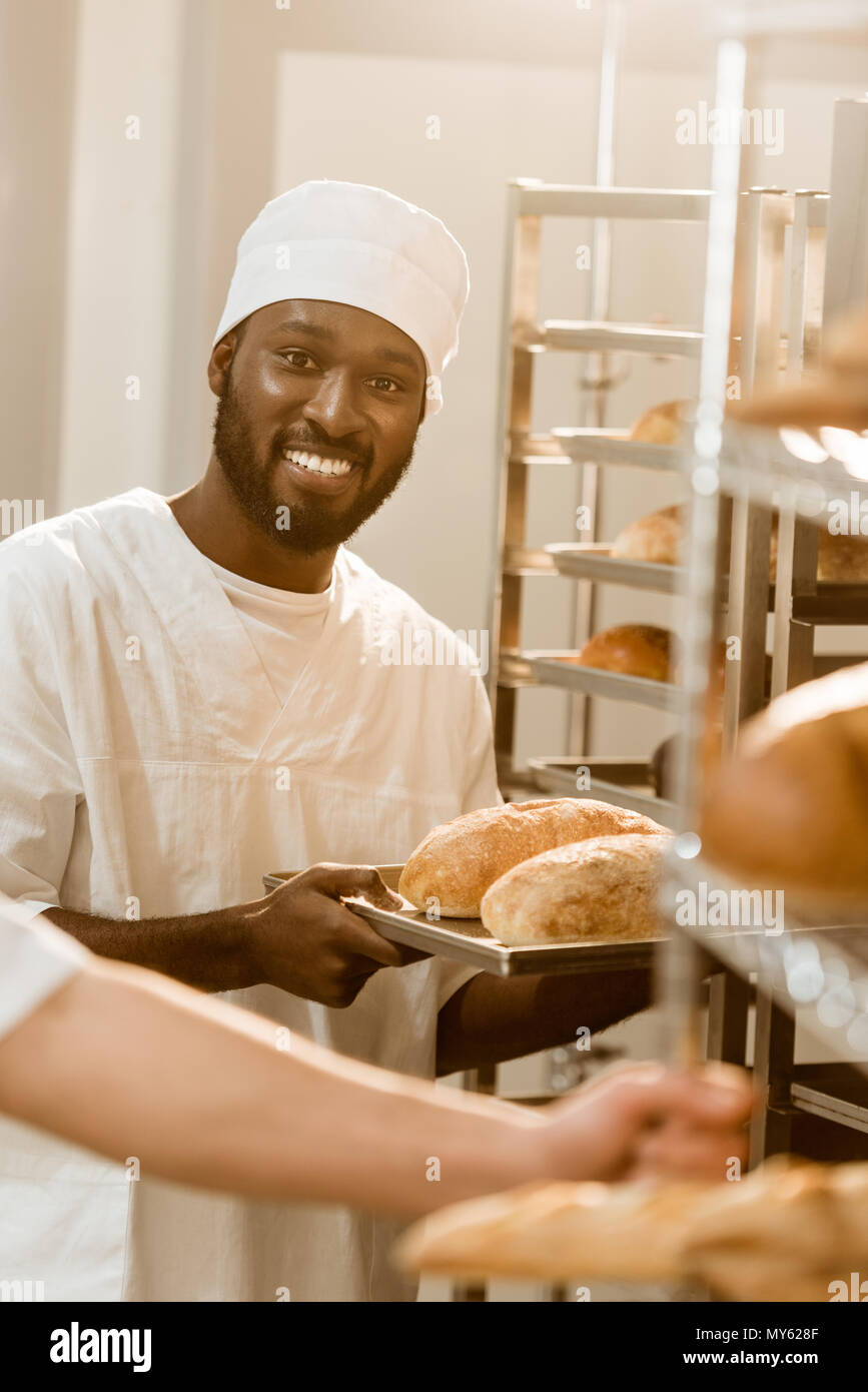 smiling african american baker holding loaves of bread on tray at baking manufacture Stock Photo