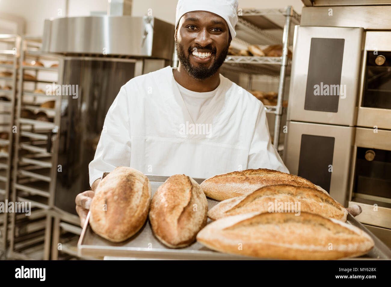 handsome african american baker with tray of fresh loaves of bread on baking manufacture Stock Photo