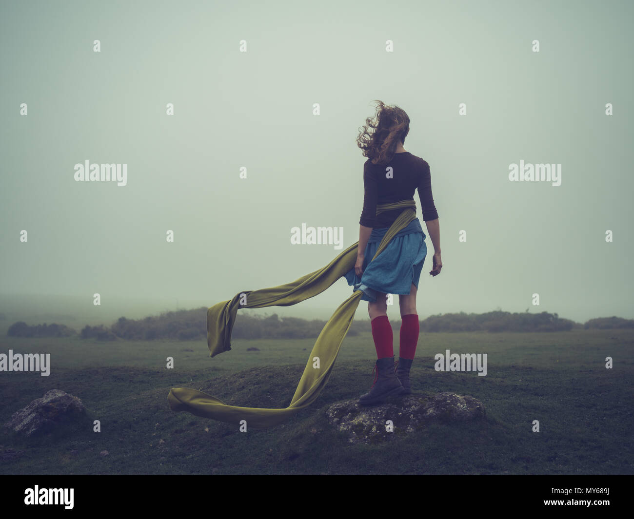 A young woman with a long scarf blowing in the wind is standing on the moor in the fog Stock Photo