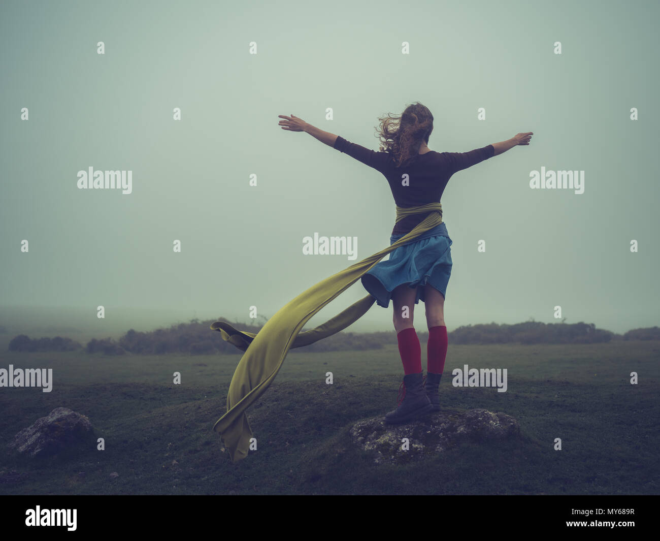 A young woman with a long scarf blowing in the wind is standing on the moor in the fog and raising her arms Stock Photo