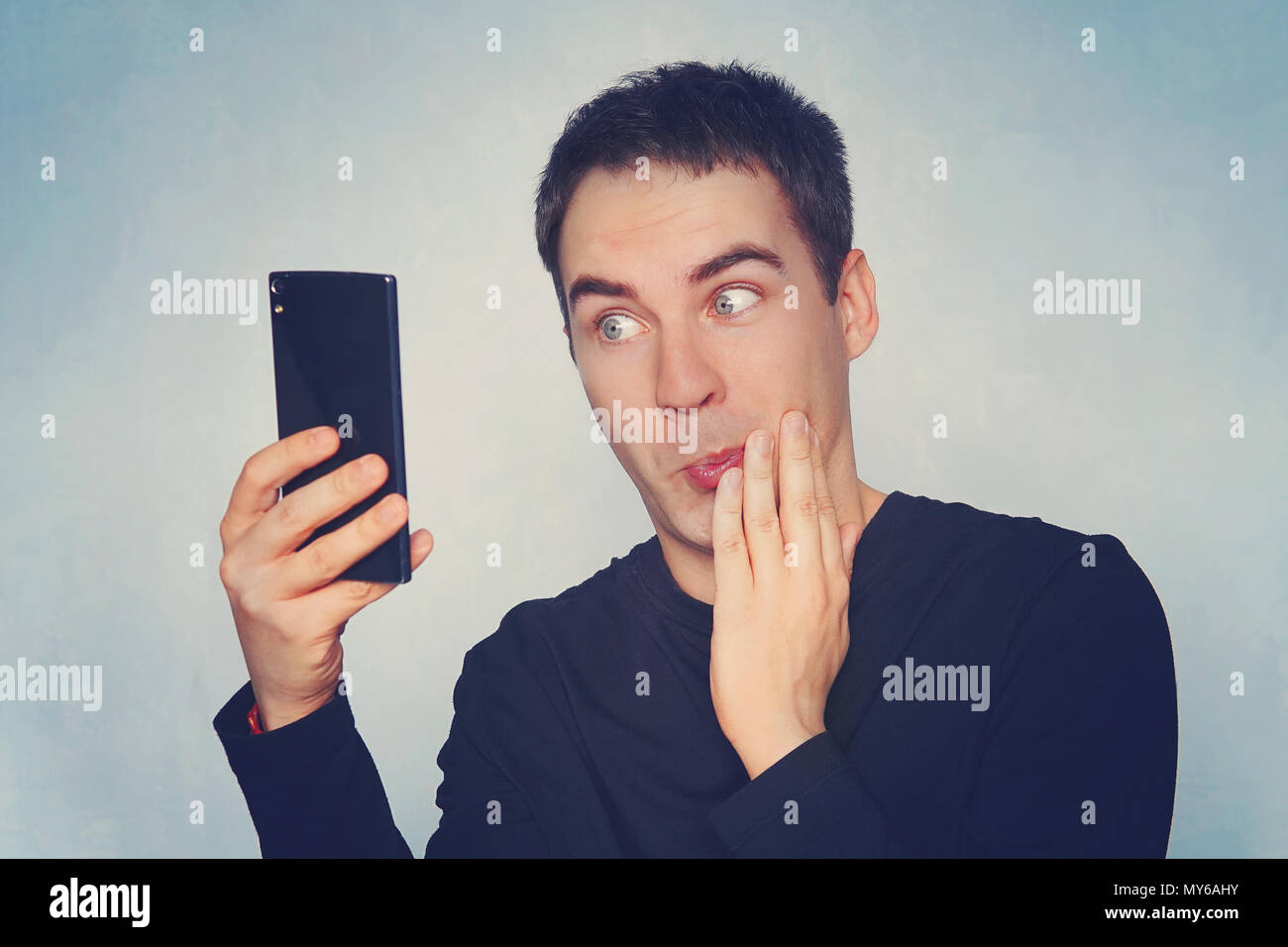 Closeup portrait of handsome young man shocked surprised, open mouth and eyes, by what he sees on his cell phone, on blue background. Negative human e Stock Photo