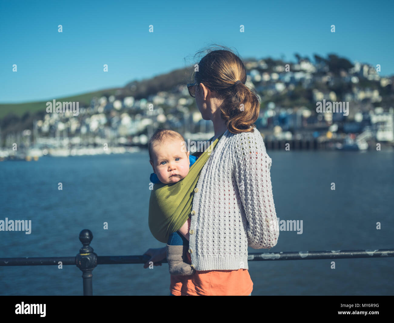 A young mother is standing by the river in a small village with her baby in a sling Stock Photo