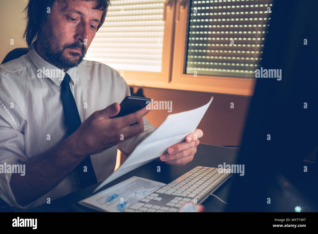 Businessman responding to a SMS text message on mobile phone while reading business letter in dark office interior, selective focus Stock Photo