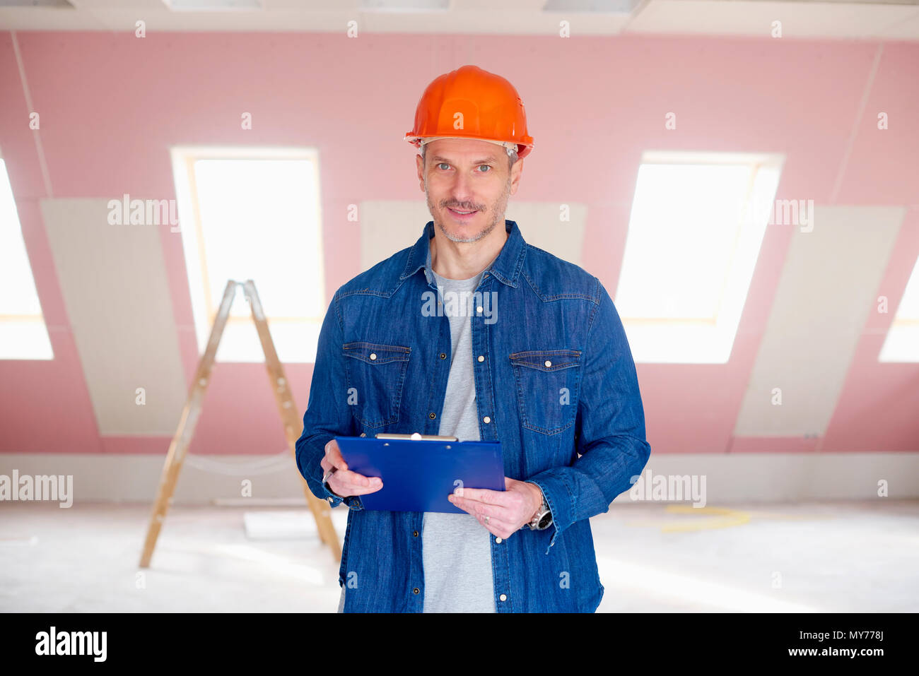 Middle aged handyman wearing hardhat and holding clipboard in his hand while standing at construction site. Stock Photo