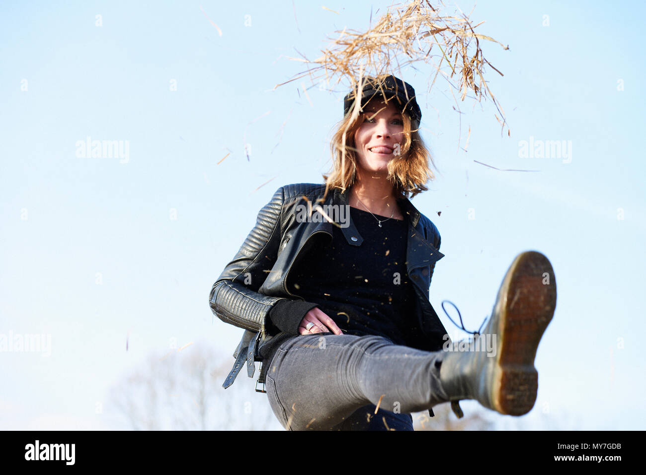 Woman in cap kicking up straw against blue sky Stock Photo