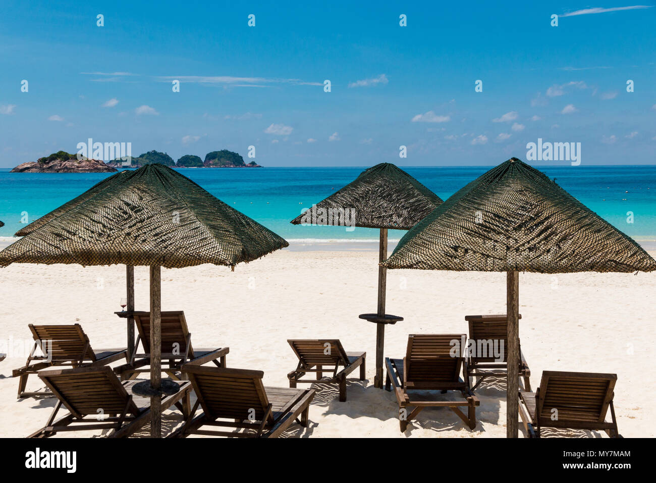 Typical holiday beach scenery with wooden sun loungers under the shade of wooden umbrellas with thatched roofs in the middle of powdery white sand. Stock Photo