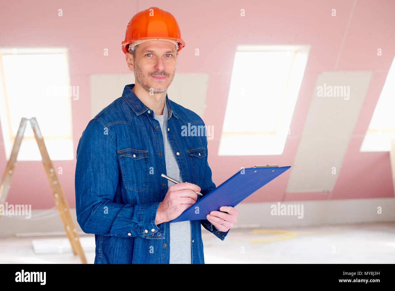 Middle aged handyman wearing hardhat and holding clipboard in his hand while standing at construction site. Stock Photo