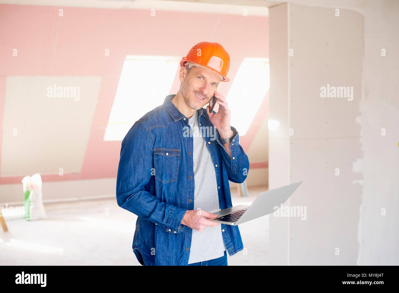 Smiling construction man making call and using his laptop while standing at construction site. Stock Photo