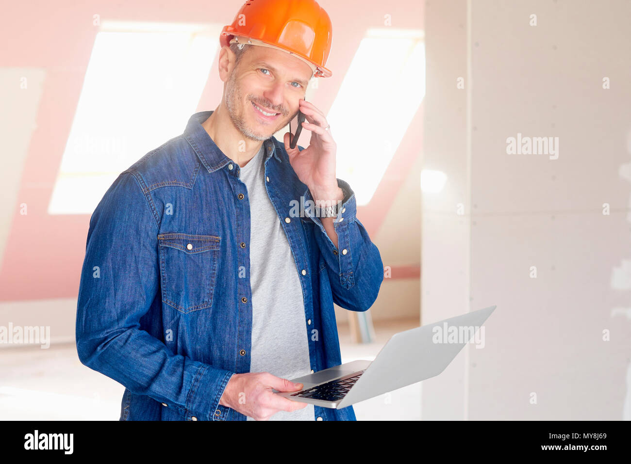 Smiling construction man making call and using his laptop while standing at construction site. Stock Photo