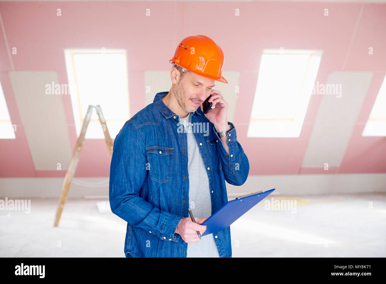 Middle aged handyman wearing hardhat and holding clipboard in his hand while while talking with somebody on mobile phone. Stock Photo