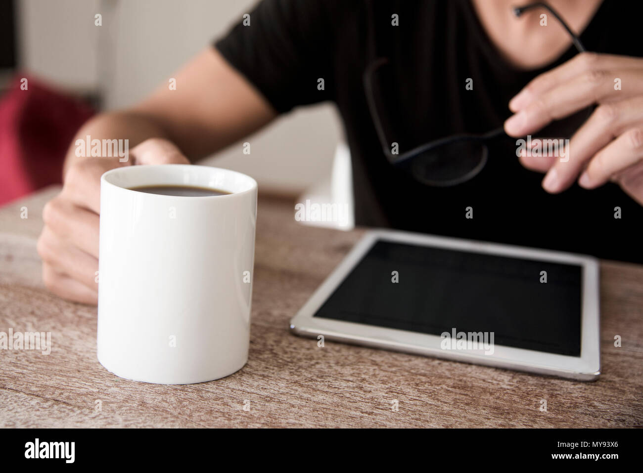 closeup of a young caucasian man, sitting at a table, using a tablet with a cup of coffee in one hand and a pair of eyeglasses in the other hand Stock Photo