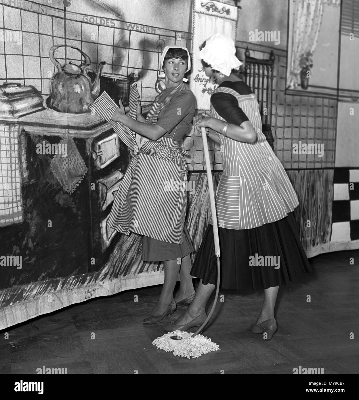 Two ladies in fashionable aprons, one with integrated potholders, in front of the picture of an old-fashioned kitchen. At the exhibition 'Du und deine Welt' of the working group of Hamburg women's organisations in the halls of Planten un Blomen in August 1955, the Master School of Fashion shows excerpts from the life of women in front of nine different pictures. | usage worldwide Stock Photo