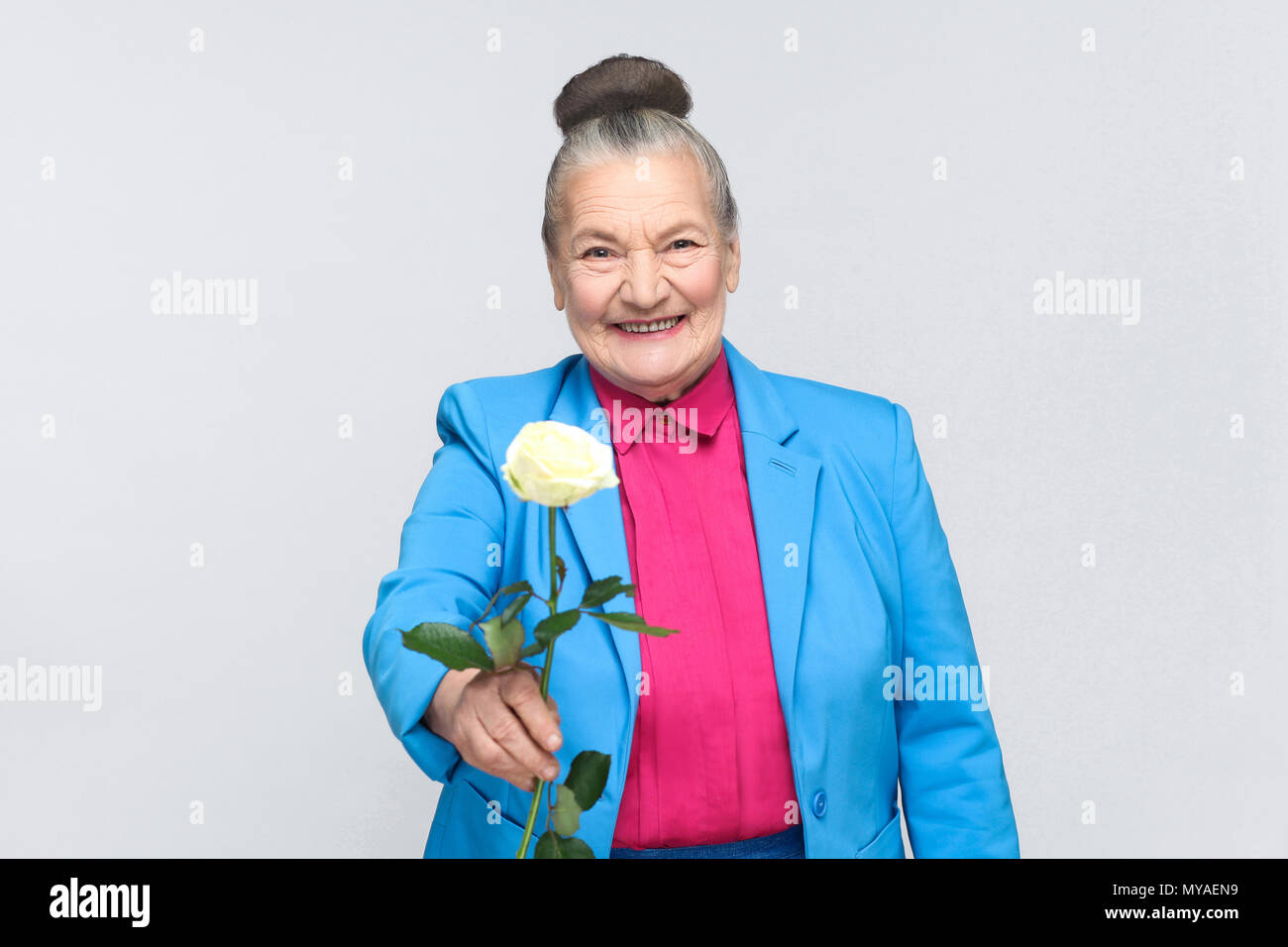 Happiness old woman present for you white rose. handsome expressive grandmother with light blue suit and pink shirt standing with collected bun gray h Stock Photo