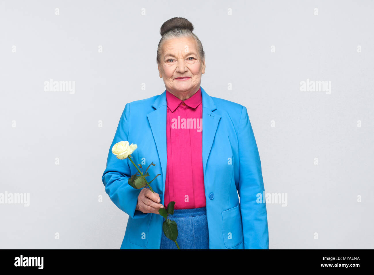 Cute old woman with white rose. Emotion and feelings concept. expressive grandmother with light blue suit and pink shirt standing with collected bun g Stock Photo