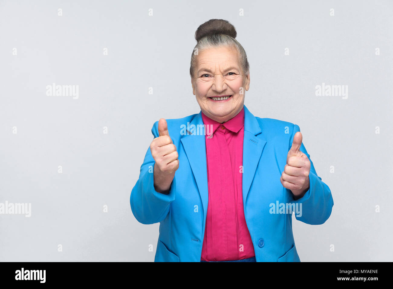 Aged woman toothy smiling with like sign. Emotion and feelings concept. expressive grandmother with light blue suit and pink shirt, collected bun gray Stock Photo