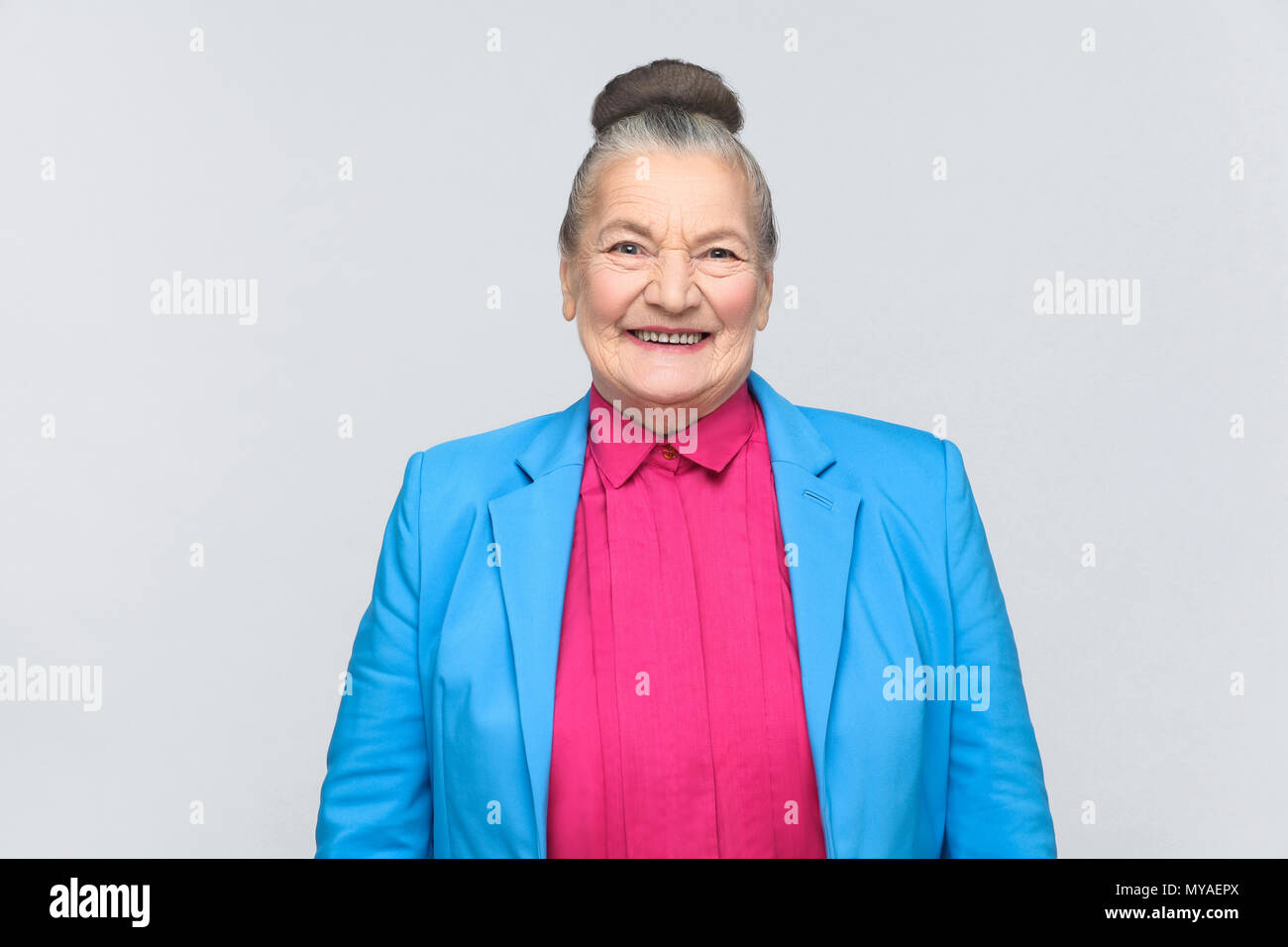Cute woman toothy smiling at camera. Emotion and feelings Portrait of expressive grandmother with blue suit standing with collected bun gray hairstyle Stock Photo