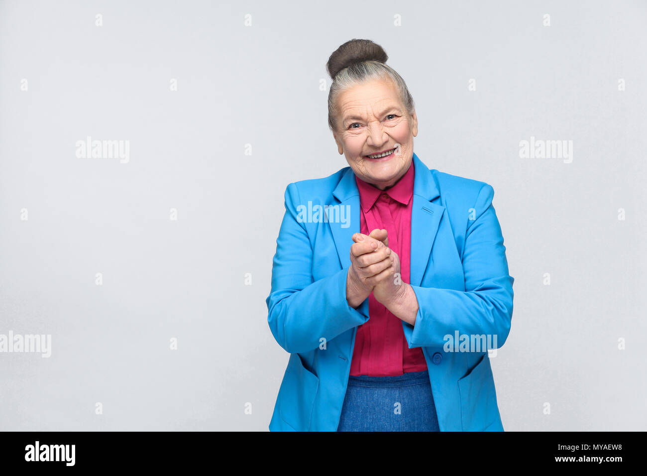 happy aged woman toothy smiling. Emotion and feelings, expressive grandmother with light blue suit and pink shirt standing with collected bun gray hai Stock Photo