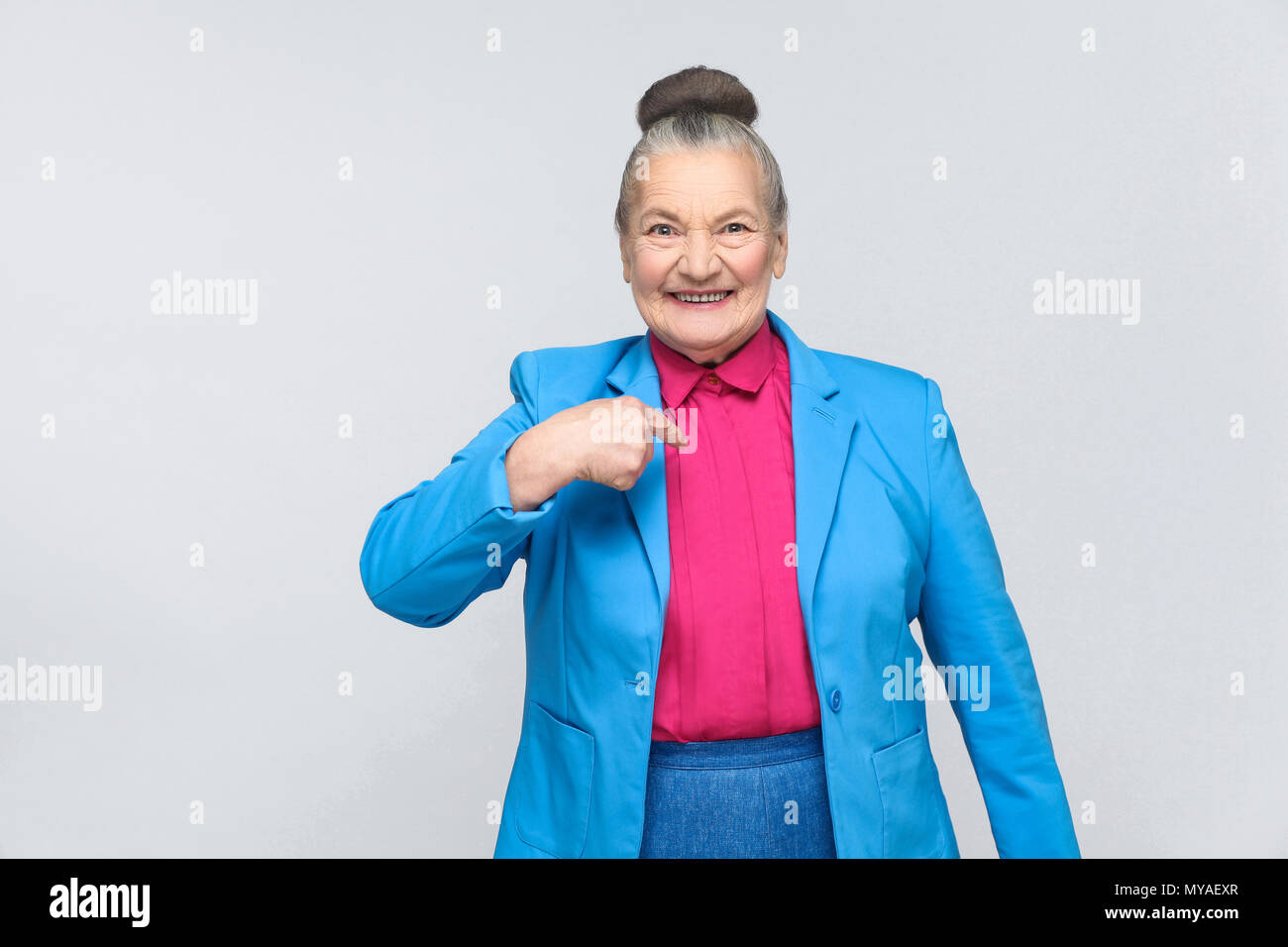 Aged woman pointing finger and toothy smiling. Emotion and feelings concept. Portrait of handsome expressive grandmother standing with collected bun g Stock Photo