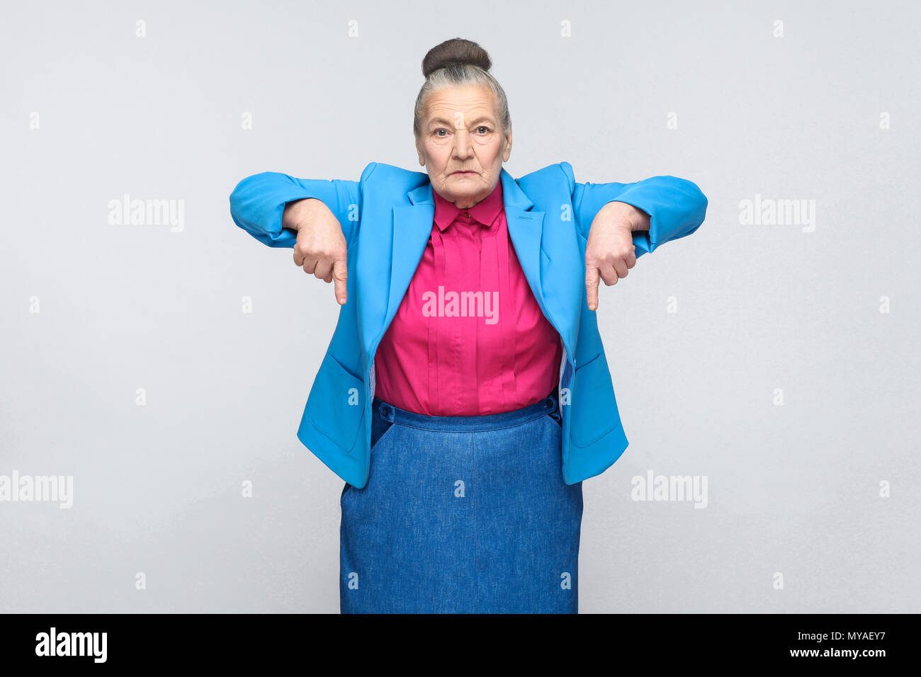 serious aged woman pointing fingers at copy space. Portrait of expressive grandmother with light blue suit and pink shirt standing with collected bun  Stock Photo