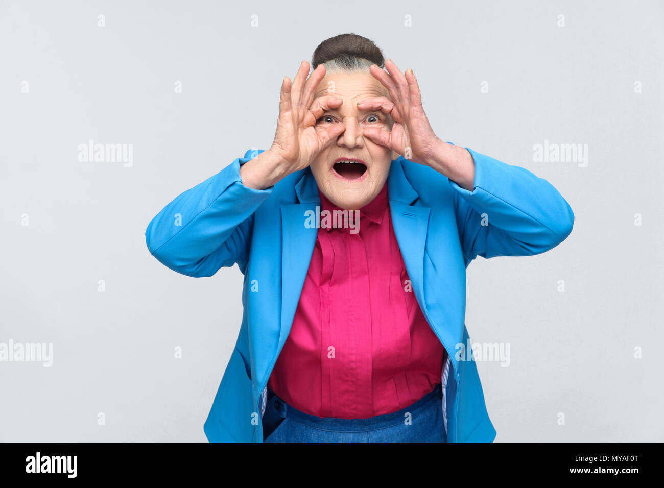 Aged grandmother with shocked face. Emotion and feelings concept. Portrait of grandmother with light blue suit and pink shirt standing with collected  Stock Photo