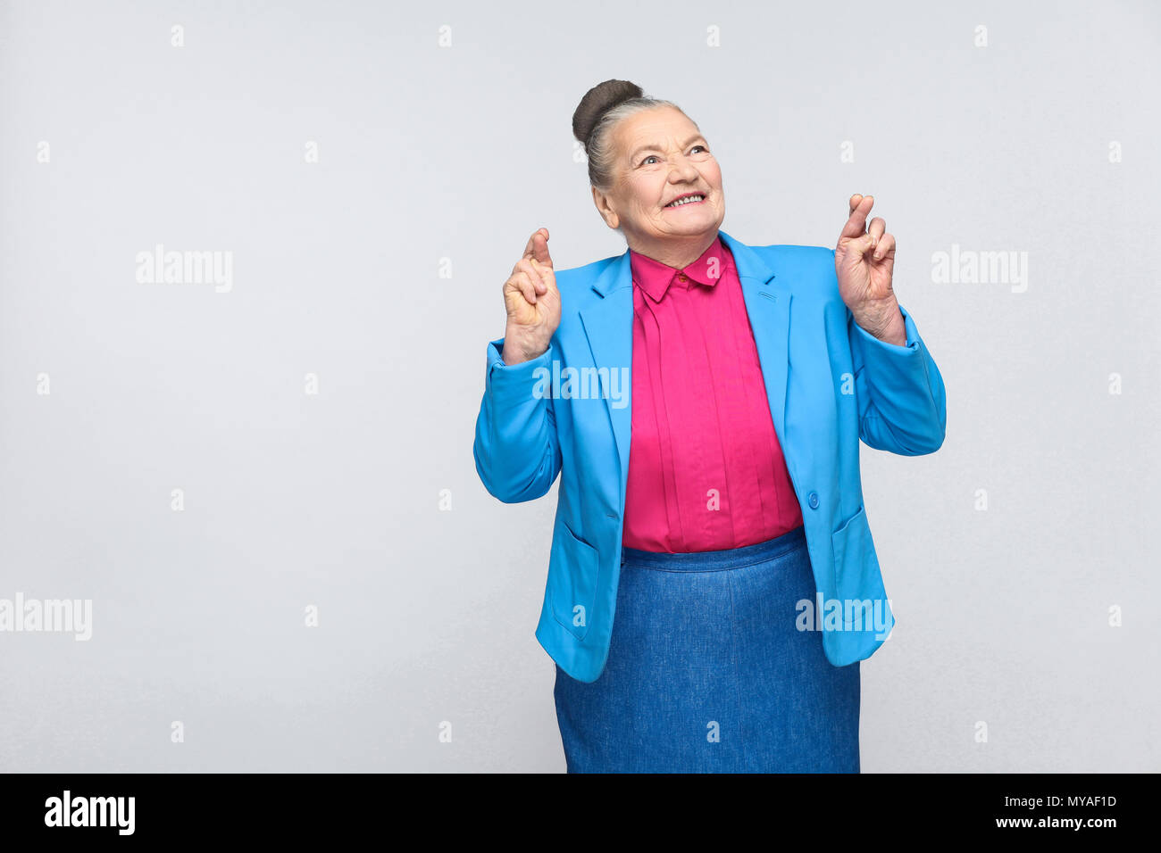 Happiness woman crossed fingers and wish. Emotion and feelings, expressive grandmother with light blue suit and pink shirt standing with collected bun Stock Photo