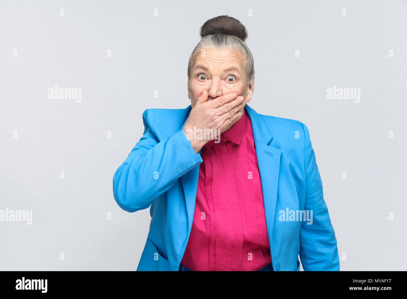 Scared aged woman closed mouth and have big eyes. Portrait of expressive grandmother with light blue suit and pink shirt standing with collected bun g Stock Photo