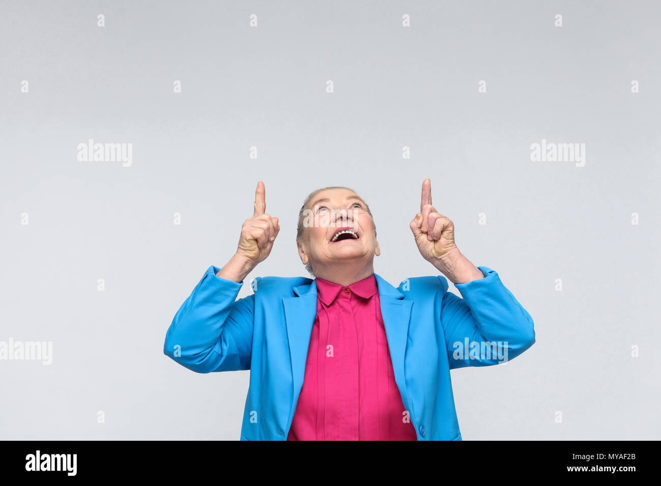 aged woman pointing finger up at copy space and toothy smiling. expressive grandmother with light blue suit and pink shirt standing with collected gra Stock Photo