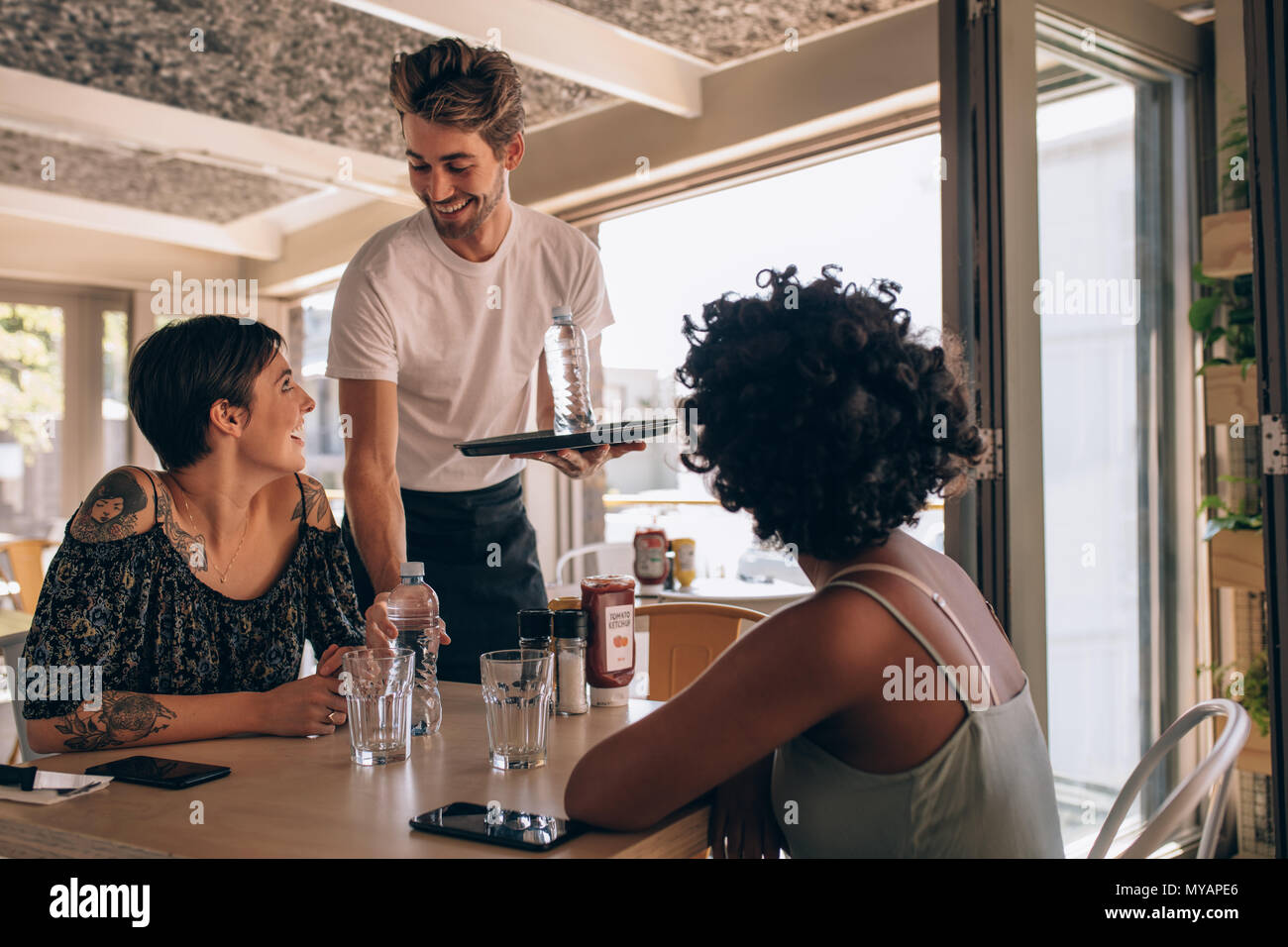 Male waiter serving water to women at cafe. Female friends at a restaurant with waiter serving water. Stock Photo