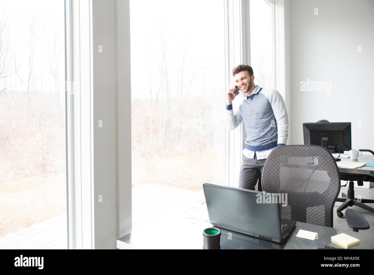 A young businessman looks out an office window while chatting on a cellphone. Stock Photo