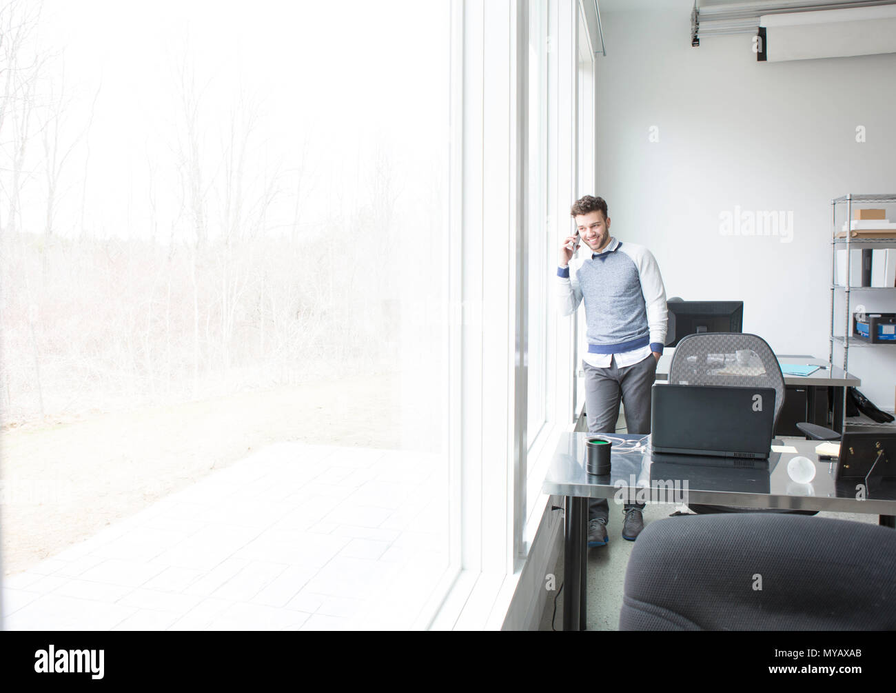 A young businessman looks out an office window while chatting on a cellphone. Stock Photo