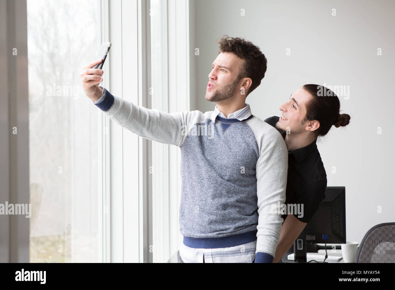 Two young businessmen make silly faces while taking selfies in a modern office. Stock Photo