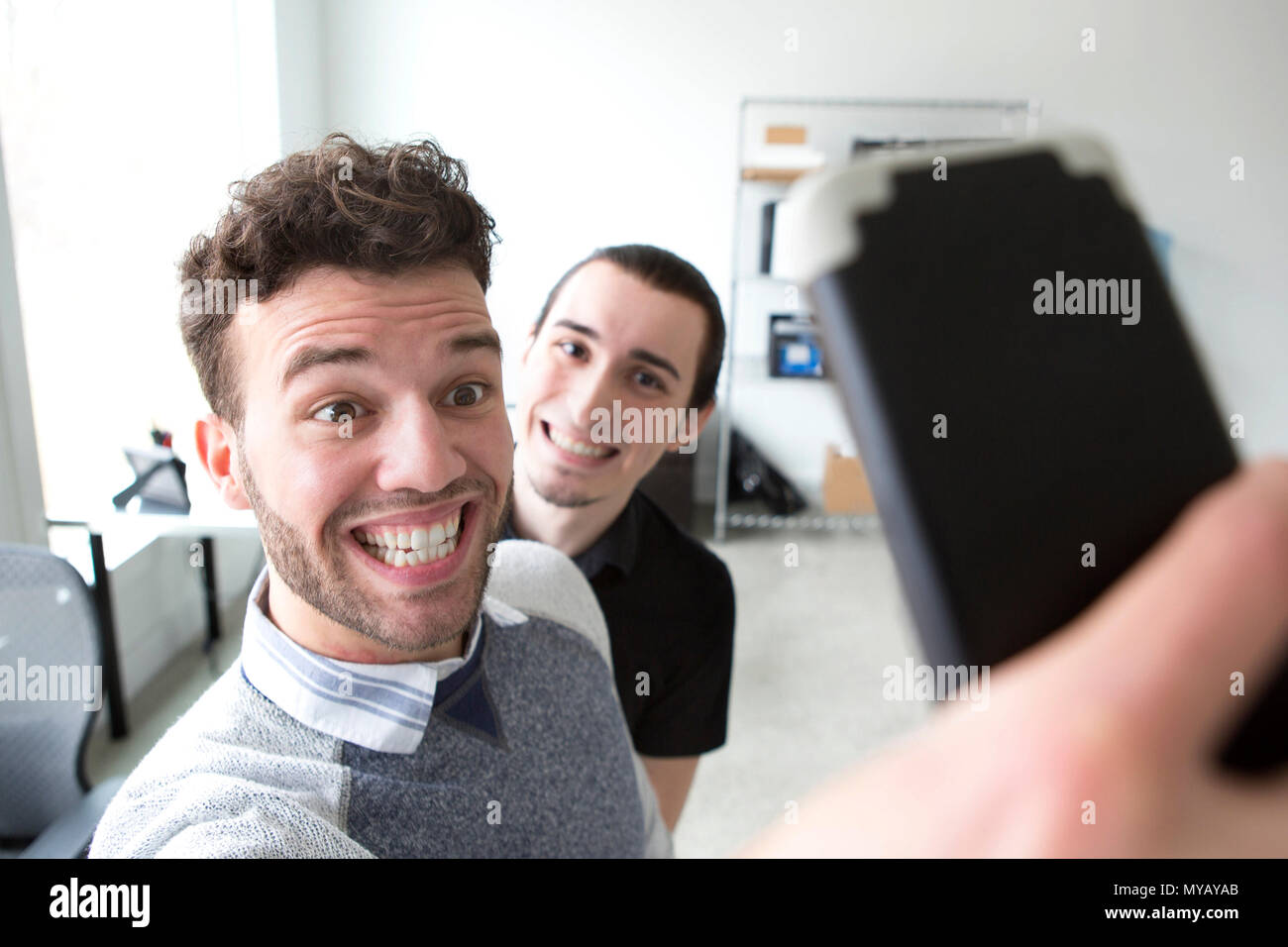 Two young businessmen make silly faces while taking selfies in a modern office. Stock Photo