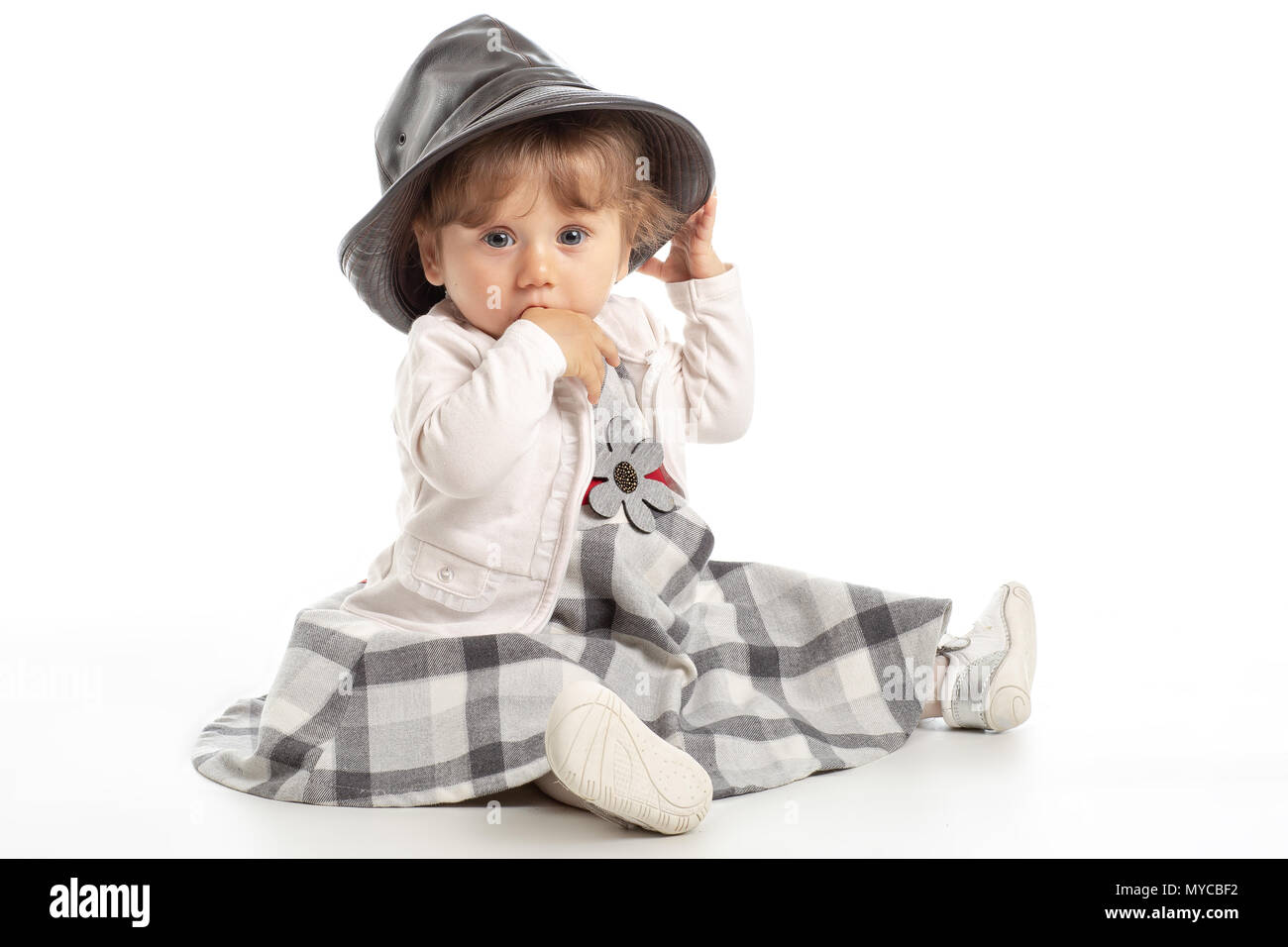 Elegant Happy baby girl 1 year old sitting on the studio floor with black leather hat. White Background. Concept Happiness. Stock Photo
