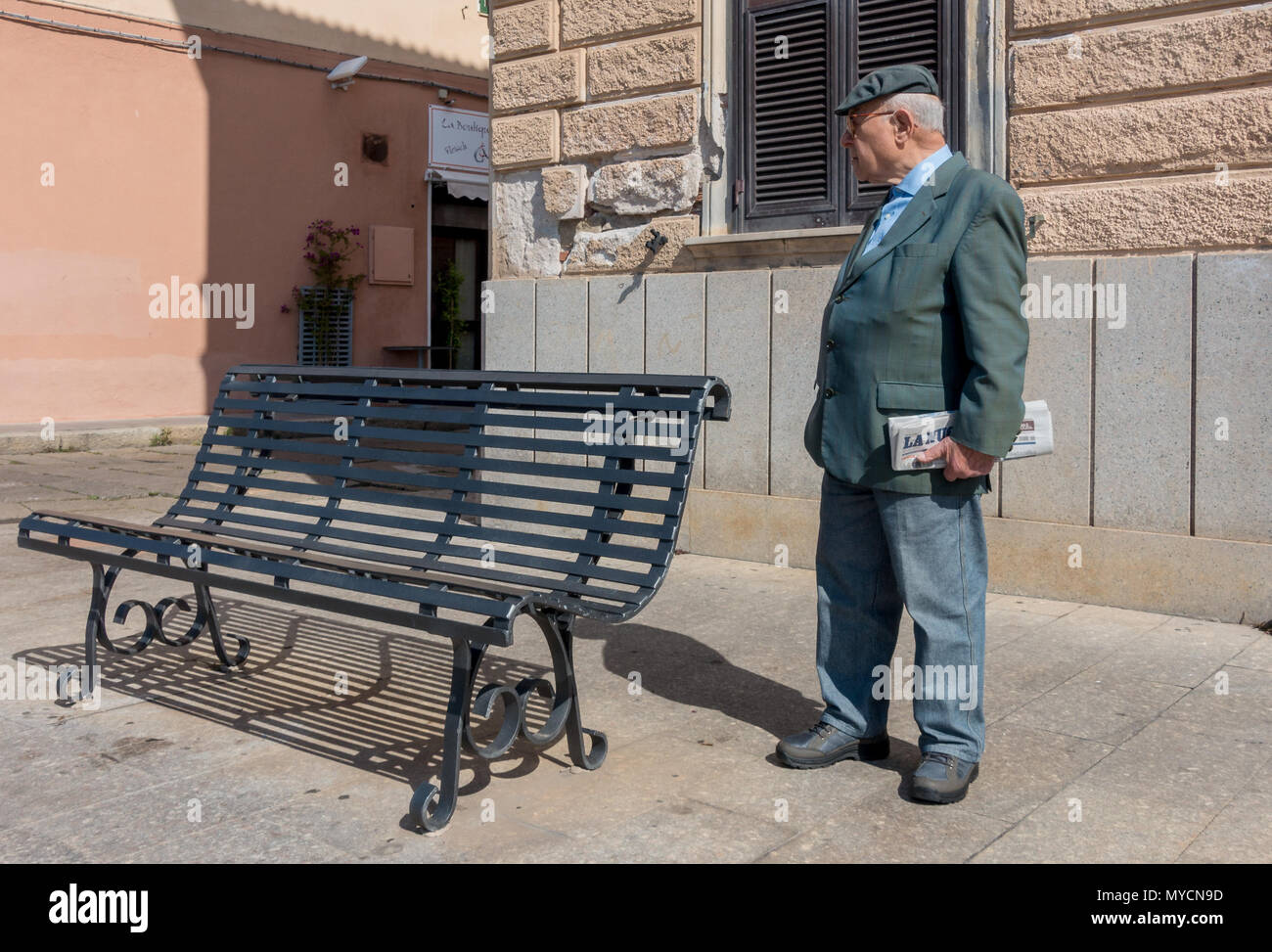 Senior Sardinian man  holding Italian newspaper with hearing aid and flat cap standing by a bench Stock Photo