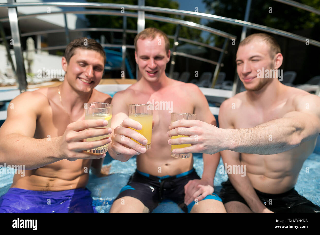 celebration, party, vacation concept. on the nosing of swimming pool that placed outdoor under the sky there is a company of three young and strong man who are clinking glasses Stock Photo