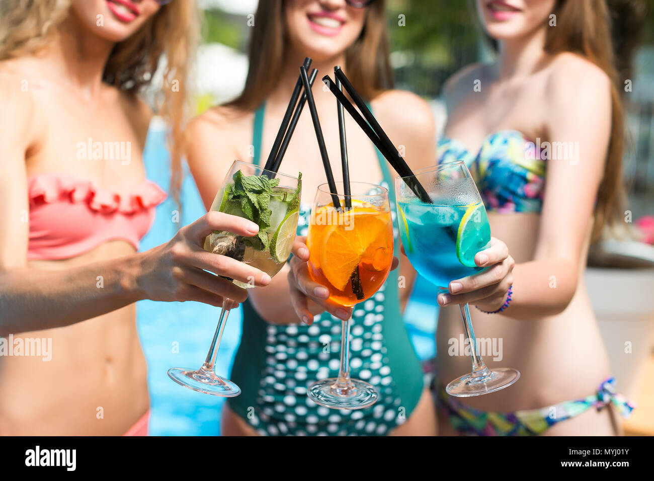 leisure time, party, celebration concept. close up of three tall glasses with different cocktails of green, orange and bright blue coloures, three young girls are holding them standing by the pool Stock Photo