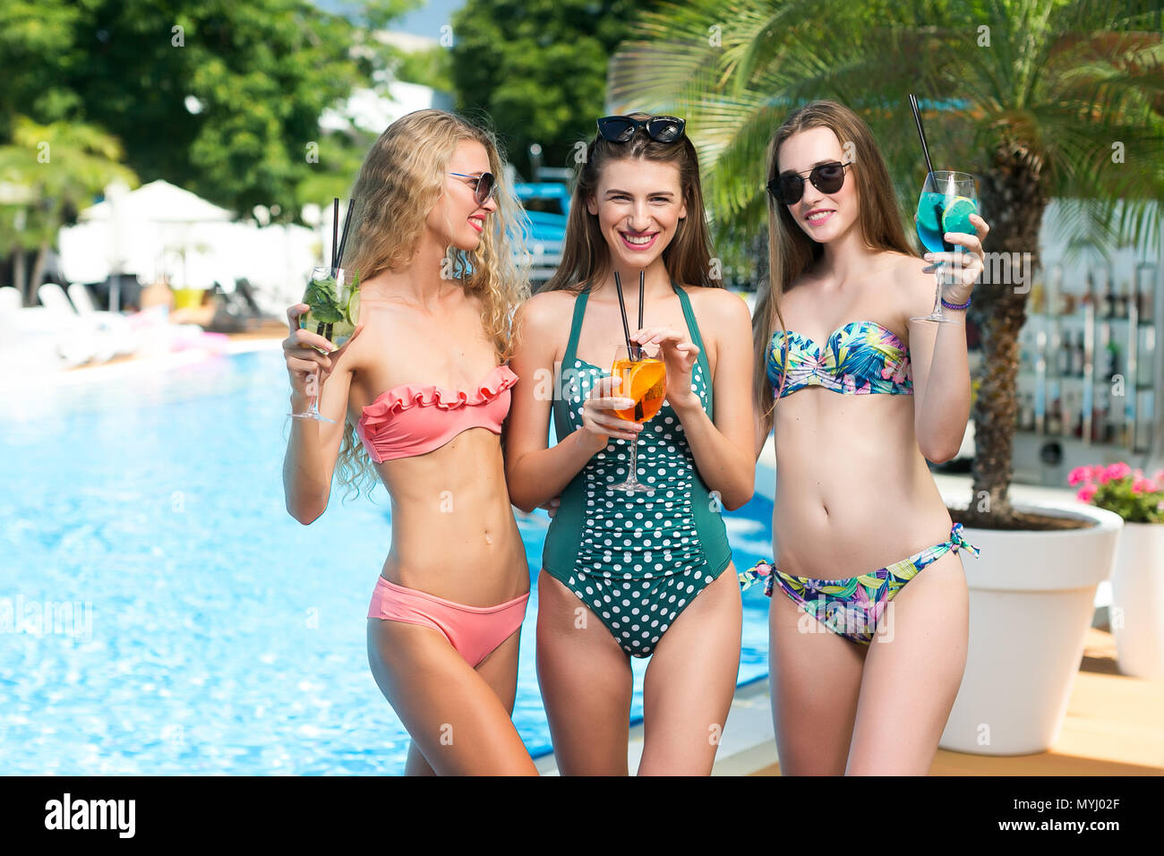 relaxation, birthday party, summer concept. by the palm tree and swimming pool with clean water that shines in the light of sun there are three slim and sexy woman who are having rest ans laughing Stock Photo