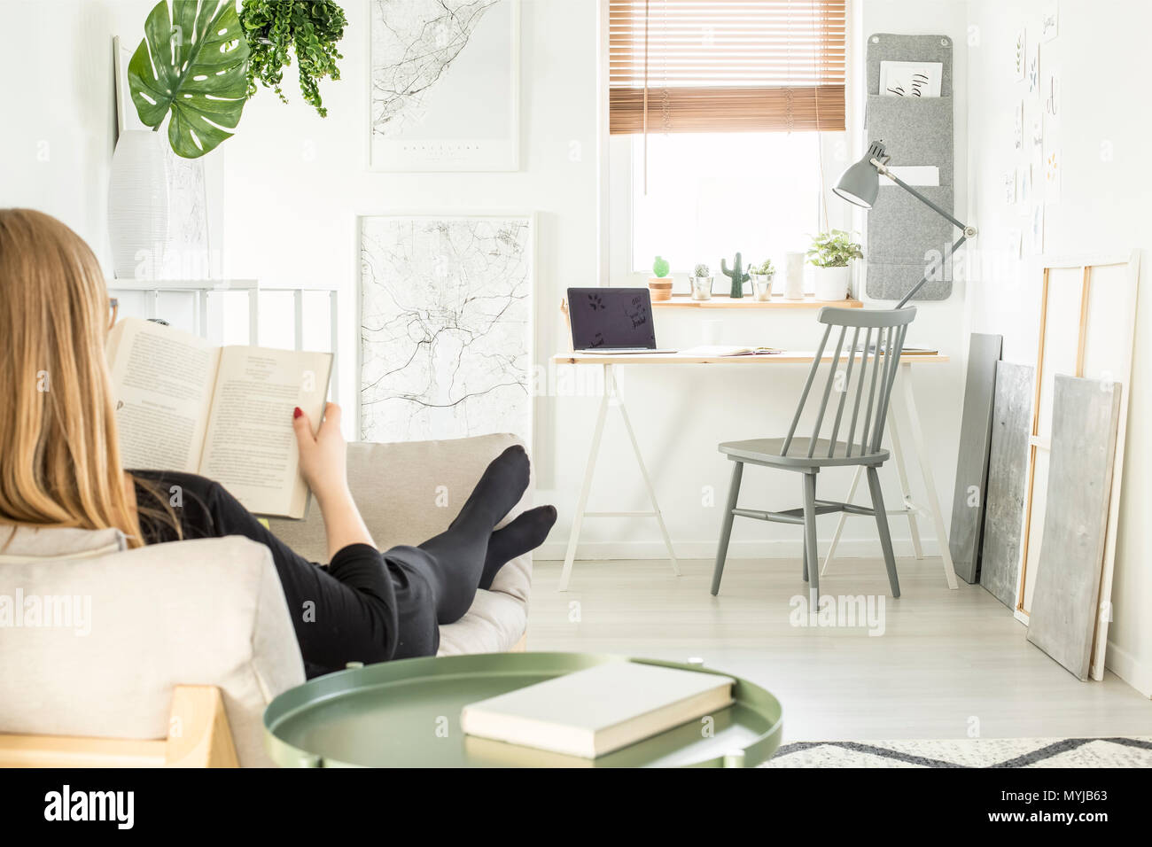 Woman reading a book on the sofa in bright home office room interior with fresh plants, lamp, laptop on desk and posters Stock Photo