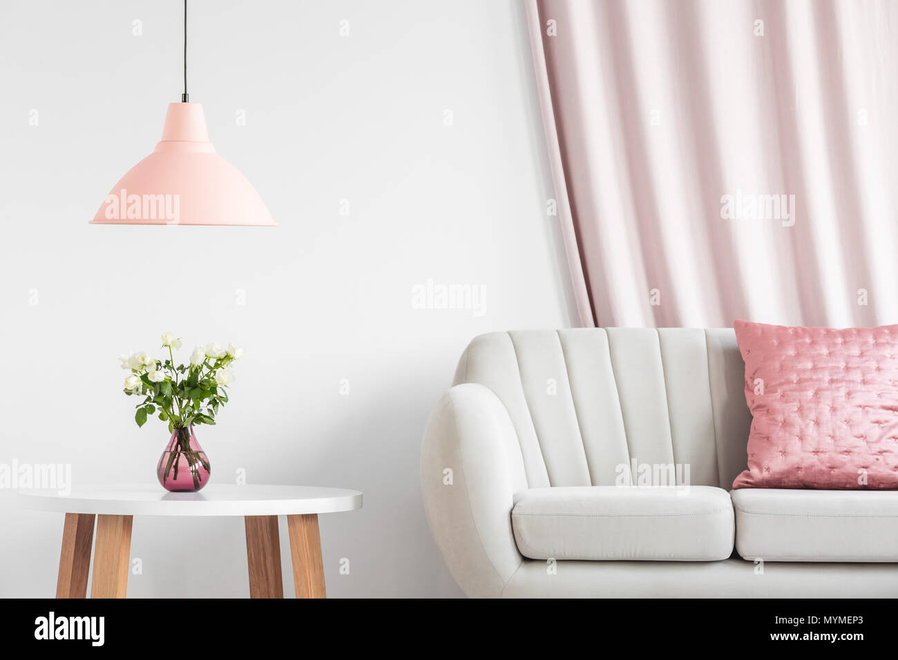 Peach lamp above wooden table with white roses in pink vase next to a sofa in living room interior Stock Photo