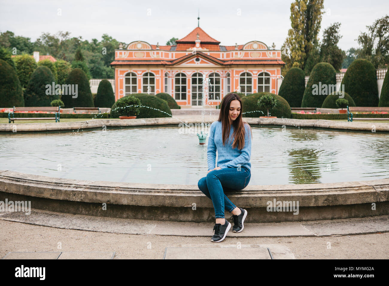 Beautiful young girl sits next to a fountain in the park and has a rest ...