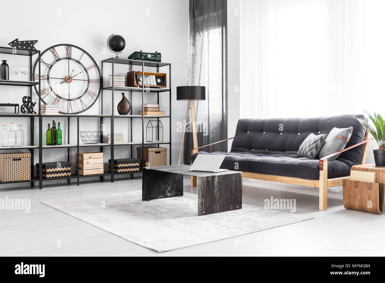 Laptop on table and wooden sofa in bright living room interior with industrial clock and bottles on a shelf Stock Photo