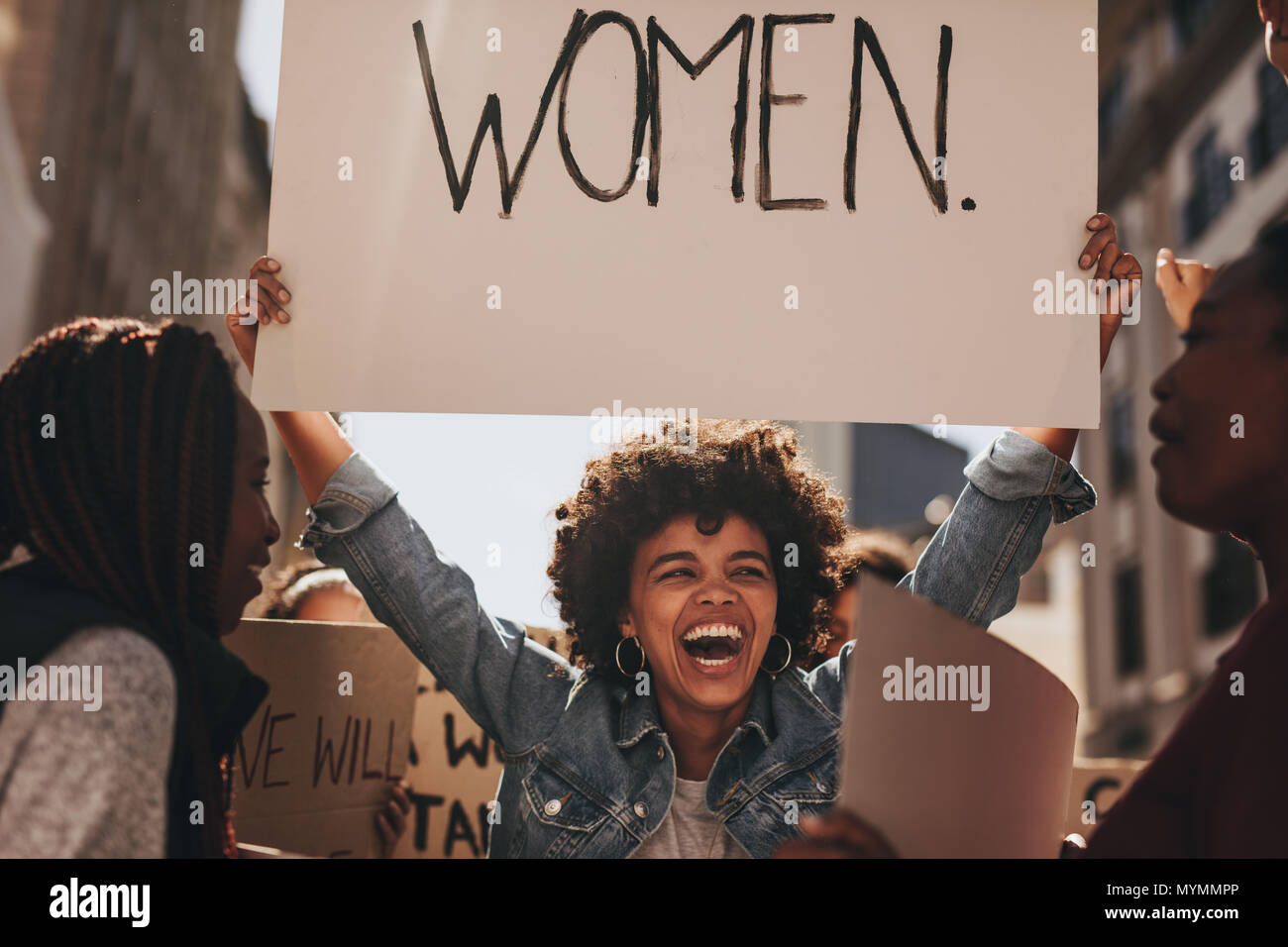 Laughing young woman holding a banner during a protest. Group of females activist protesting on road for women empowerment. Stock Photo