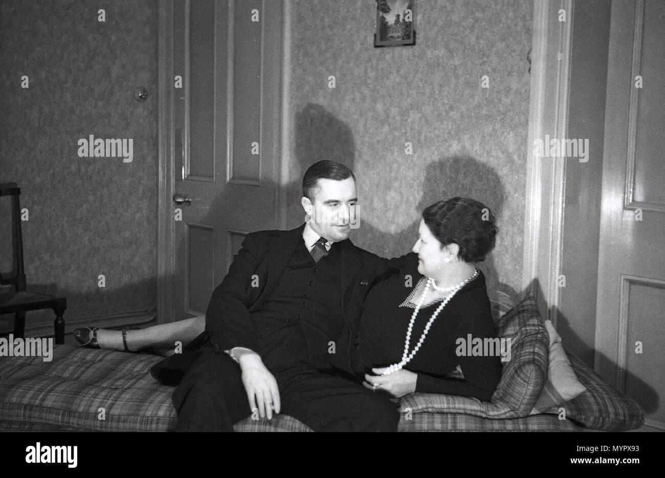 1936, historical, early evening and a well-dressed gentleman and lady relax together on a chaise longue in a front room, London, England, UK. Widely used in wealthy residences in this era, a chaise longue is an upholstered sofa in the shape of a chair but that is long engough to support the legs. Its literal translation is 'long chair'. The modern chaise longue first became popular in France in the 16th century, created by furniture craftsmen for their rich customers, for them to rest without having to go the bedroom. Stock Photo