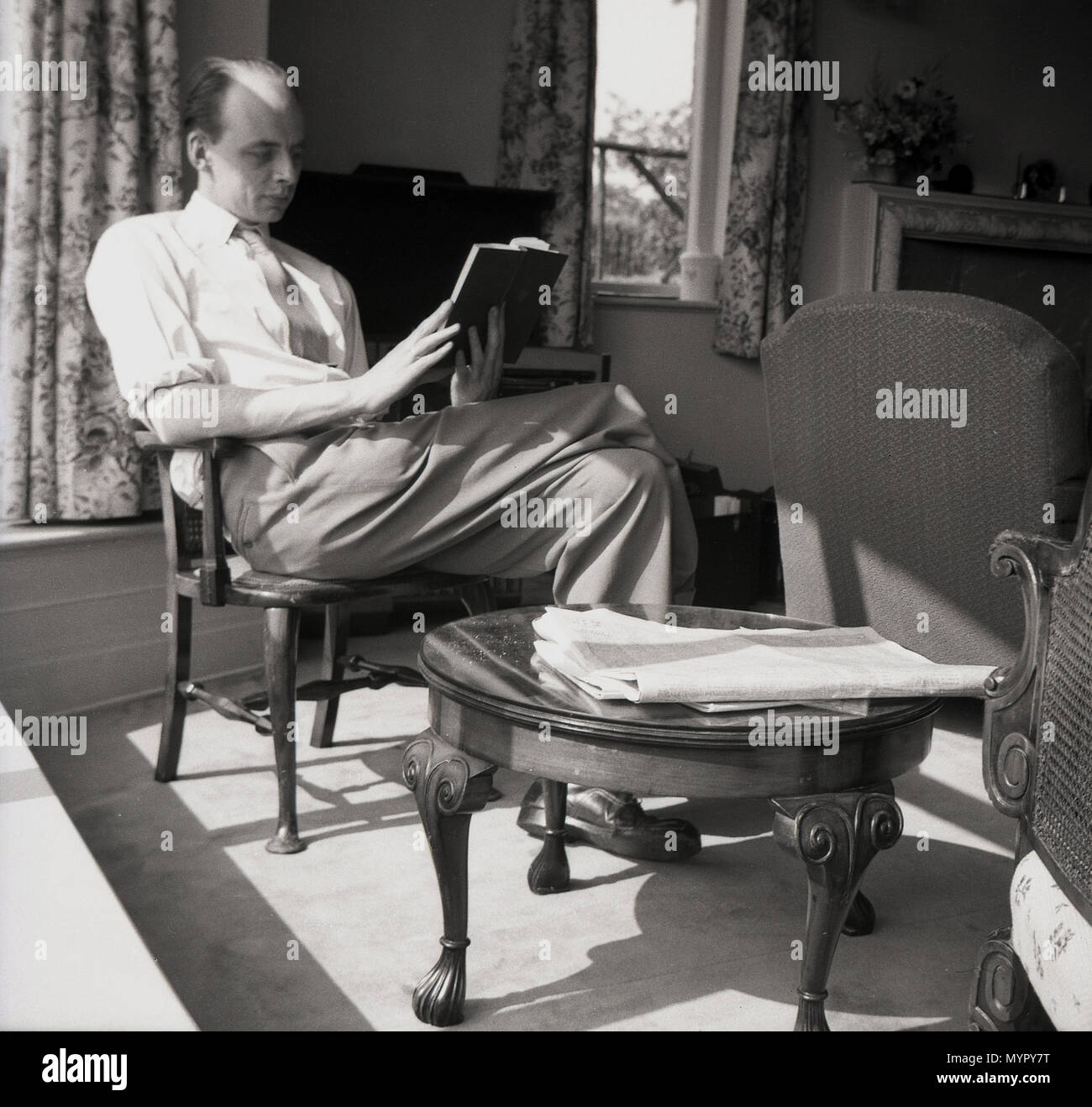 1953, July, an adult man, with an unusual hairstyle, sitting in a chair in a front room reading a book, England, UK. Stock Photo