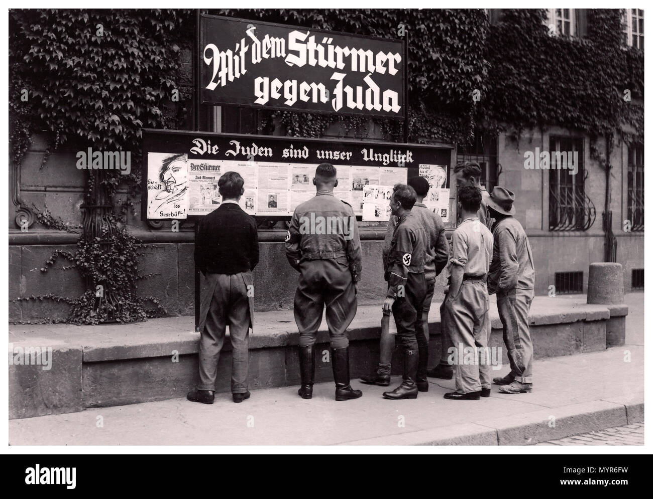 Vintage 1930’s Nazi Germany Propaganda Newspaper anti Semitic Der Stürmer (The Attacker) is posted in the street with NSDAP members and a motley dissolute group of German men reading the racist inflammatory headline which reads “The Jews are our Misfortune” City of Worms Germany 1935 Stock Photo