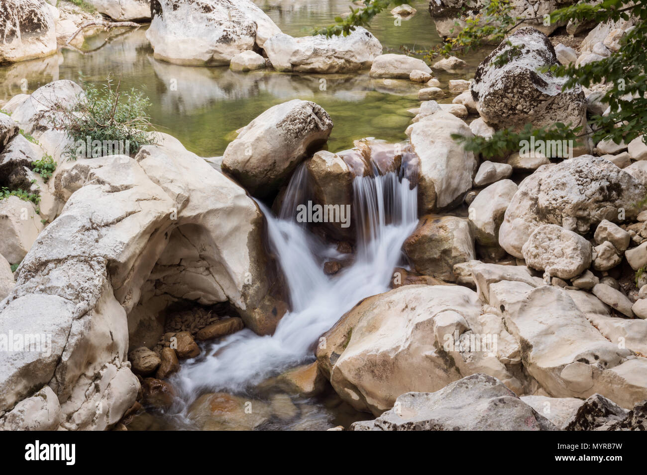 A tranquil waterfall cascades through smooth, weathered rocks into a serene pool of water. Surrounded by lush greenery and natural rock formations, Stock Photo
