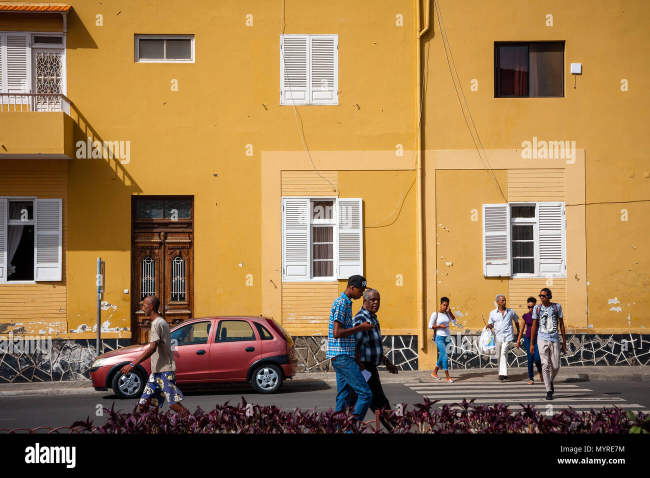 Life in Mindelo, residents walking the streets. Town architecture,  large yellow building wall MINDELO, CAPE VERDE - DECEMBER 07, 2015 Stock Photo