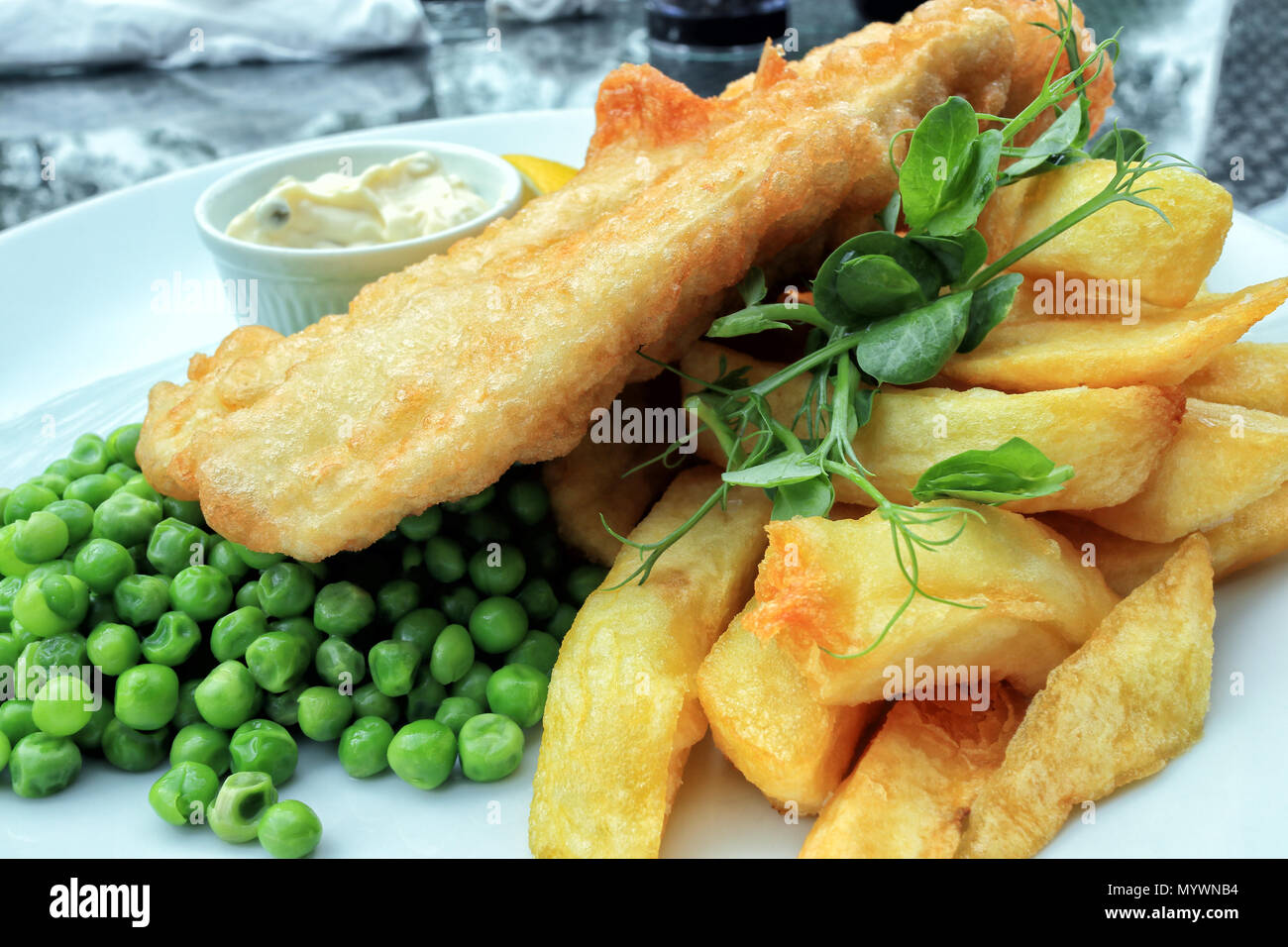 Crispy battered fish and chunky potato chips served with boiled green peas and mayonnaise, traditional English food . Stock Photo