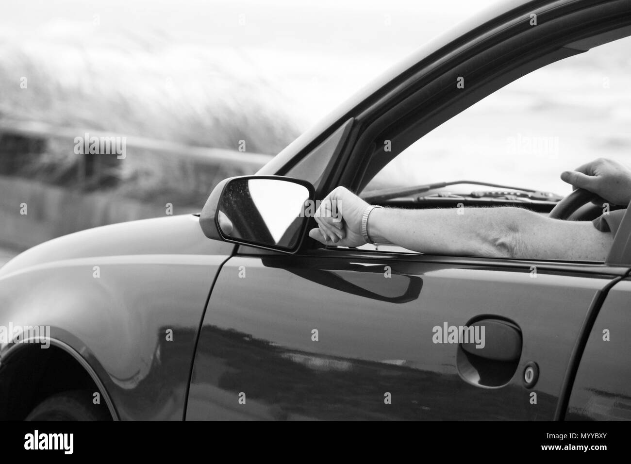 Black and White photo of someone driving a car on a road trip.  One arm on the steering wheel and the other is resting on the door with the window dow. Stock Photo