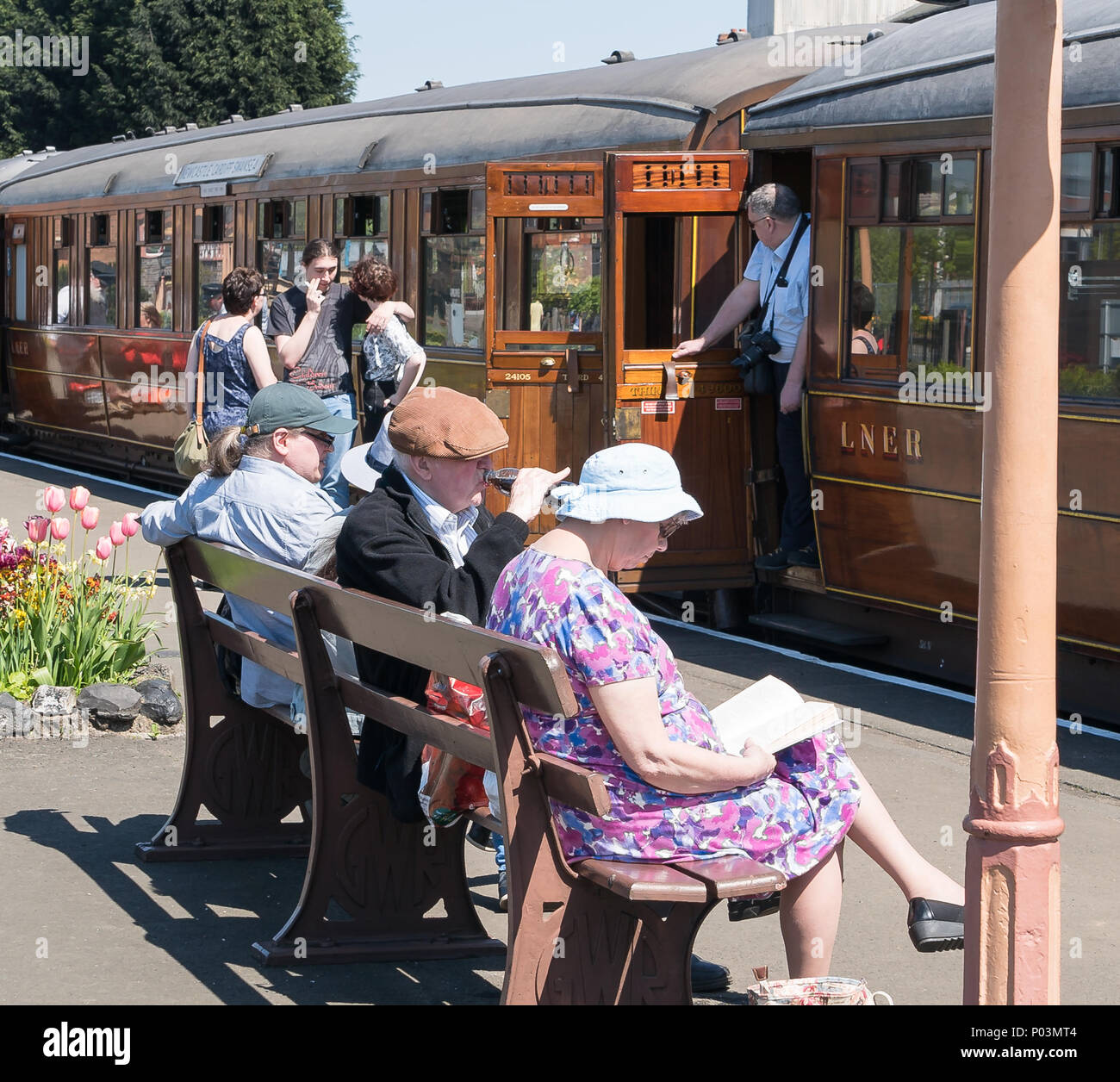 Busy platform scene at vintage railway station in summer sunshine. Passengers on & off vintage train. British folk sat on bench waiting & enjoying sun. Stock Photo